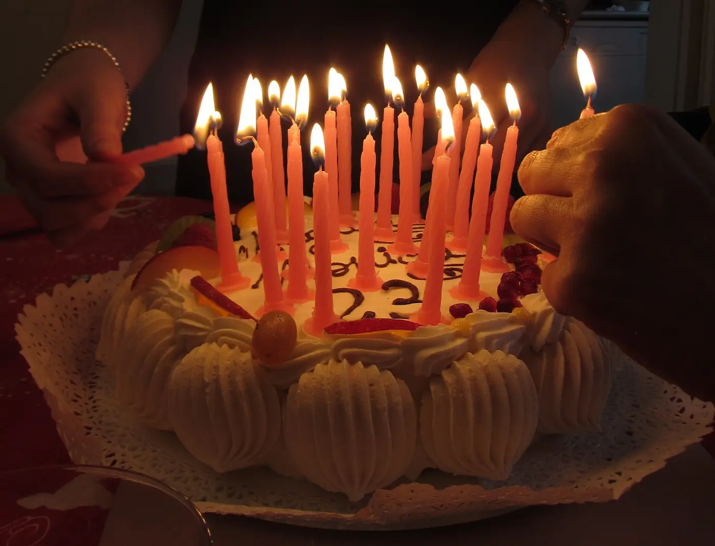 Birthday cake with a chocolate 23 and lots of lit candles, two hands lighting the last ones