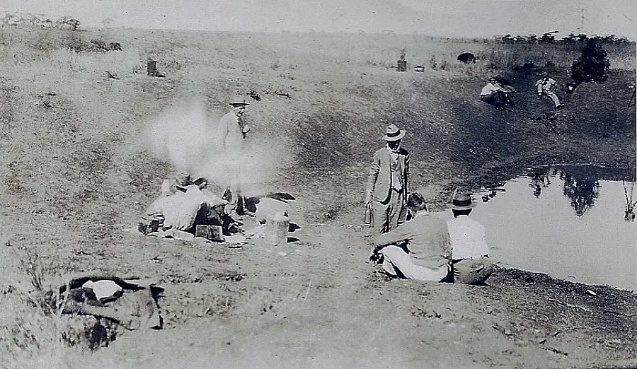 Historical photo: men in hats resting at a waterhole on dry land, more people in the background