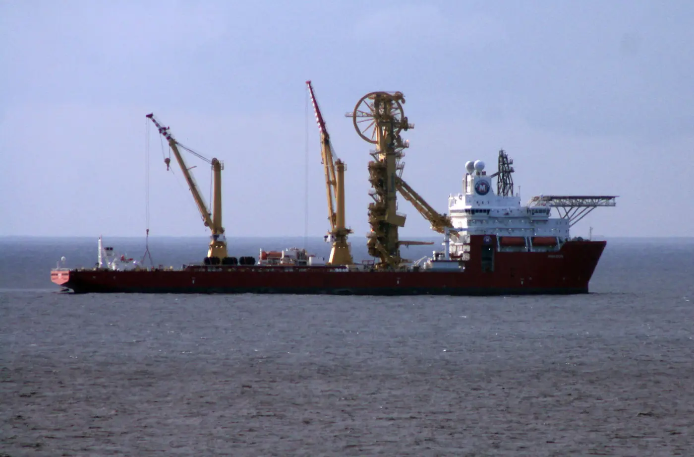 A red special-purpose ship with yellow cranes and a large cable drum at sea