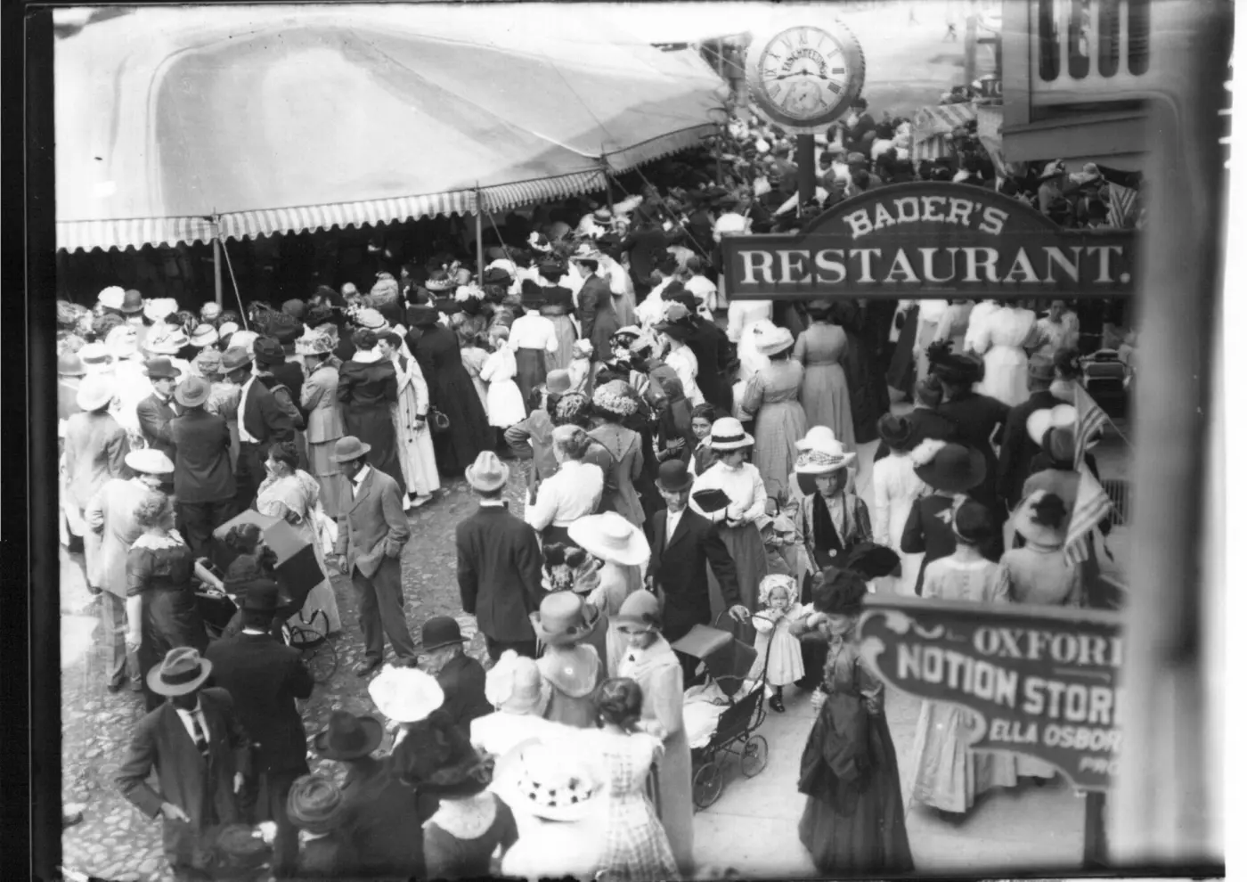 Historical photo: a dense crowd at a street fair, people in hats in front of shop signs