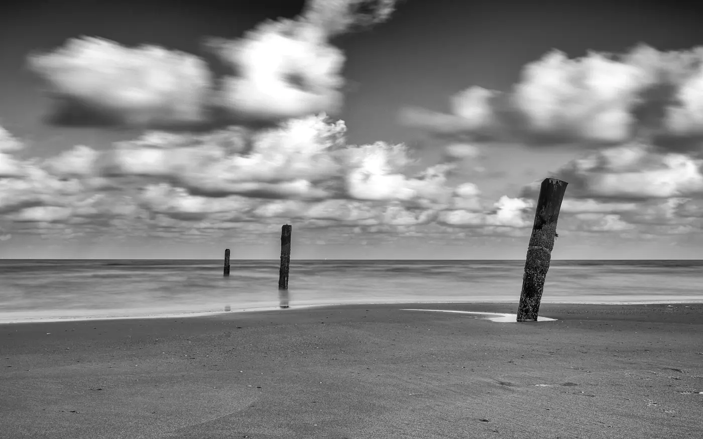 Black-and-white photo: a flat sandy beach with old wooden posts, behind it the sea and a straight horizon under clouds