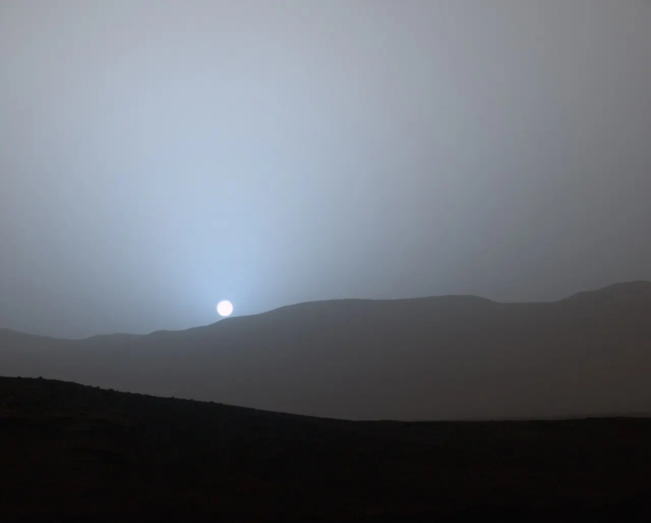 A pale, bluish glowing sun above dark hills on the Martian horizon