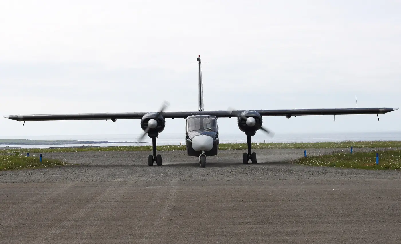 A small twin-engine aircraft on a gravel airstrip under a grey sky